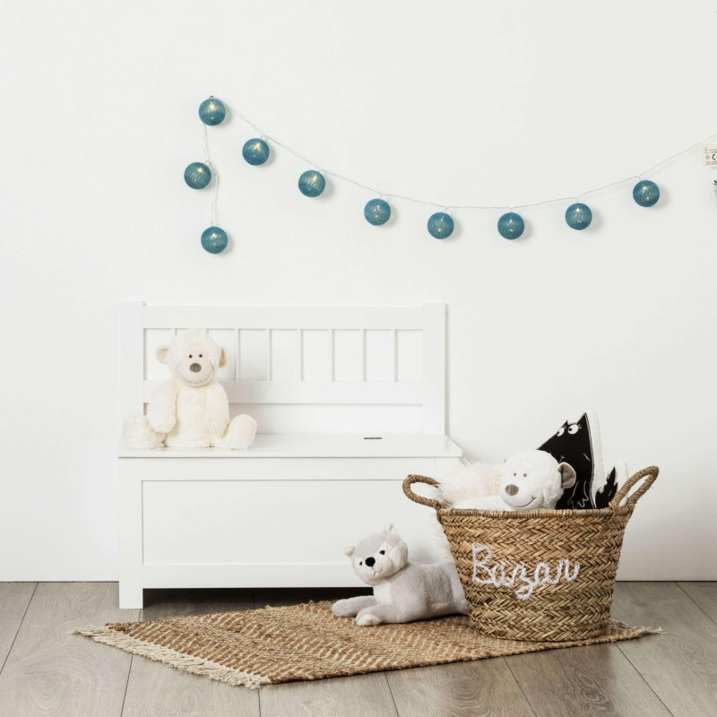 Baúl infantil blanco con respaldo decorado con juguetes y guirnalda de esferas azules en ambiente de dormitorio
