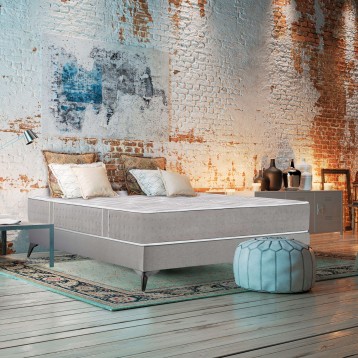 Cama tapizada gris claro con colchón blanco y almohadas decorativas en ambiente de loft con pared de ladrillo visto