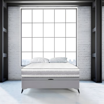 Cama tapizada gris claro con cabecero acolchado y patas metálicas en ambiente moderno junto a ventana de grid