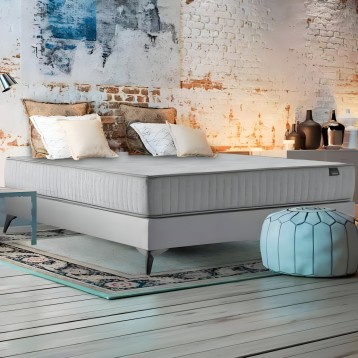 Cama con base blanca y colchón gris tapizado con almohadas decorativas en ambiente industrial con pared de ladrillo visto
