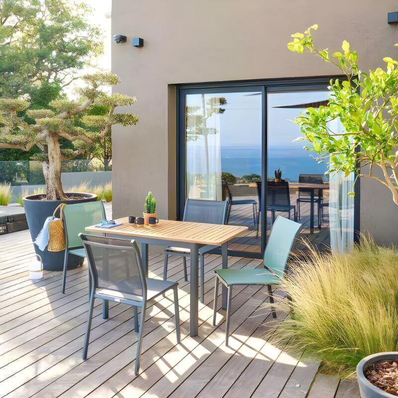 Conjunto de comedor de exterior con mesa rectangular de madera y sillas de respaldo azul turquesa en terraza con vistas al mar
